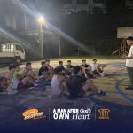 Programs 41 A man speaking to a group of young people sitting on a basketball court at night.