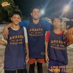 Programs 33 Three young boys in basketball jerseys smiling at night outdoors.