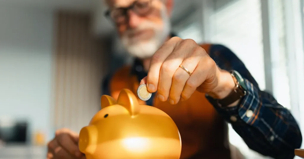 Elderly man putting coin into a golden piggy bank for savings.